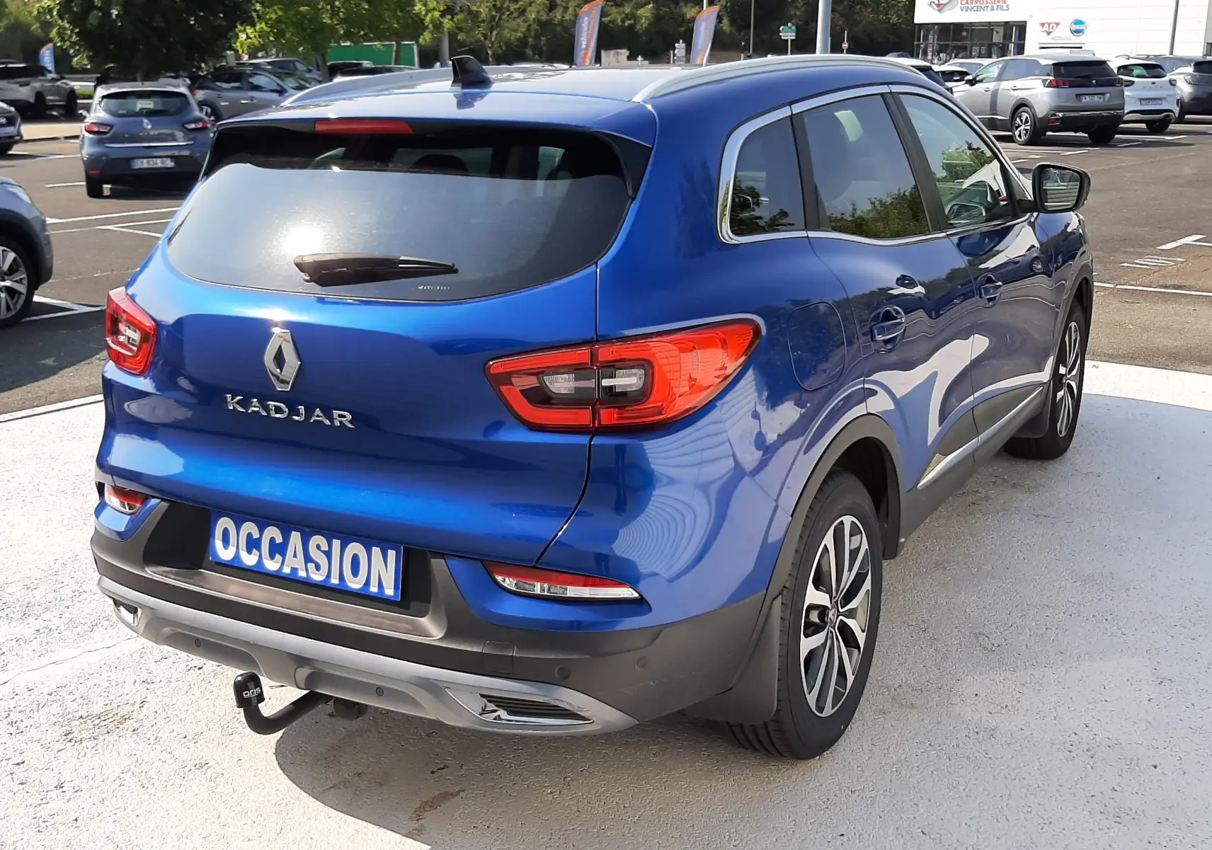 Vue 3/4 arrière droite d'un Renault Kadjar bleu Iron 2020 avec attelage et plaque "OCCASION" visible.