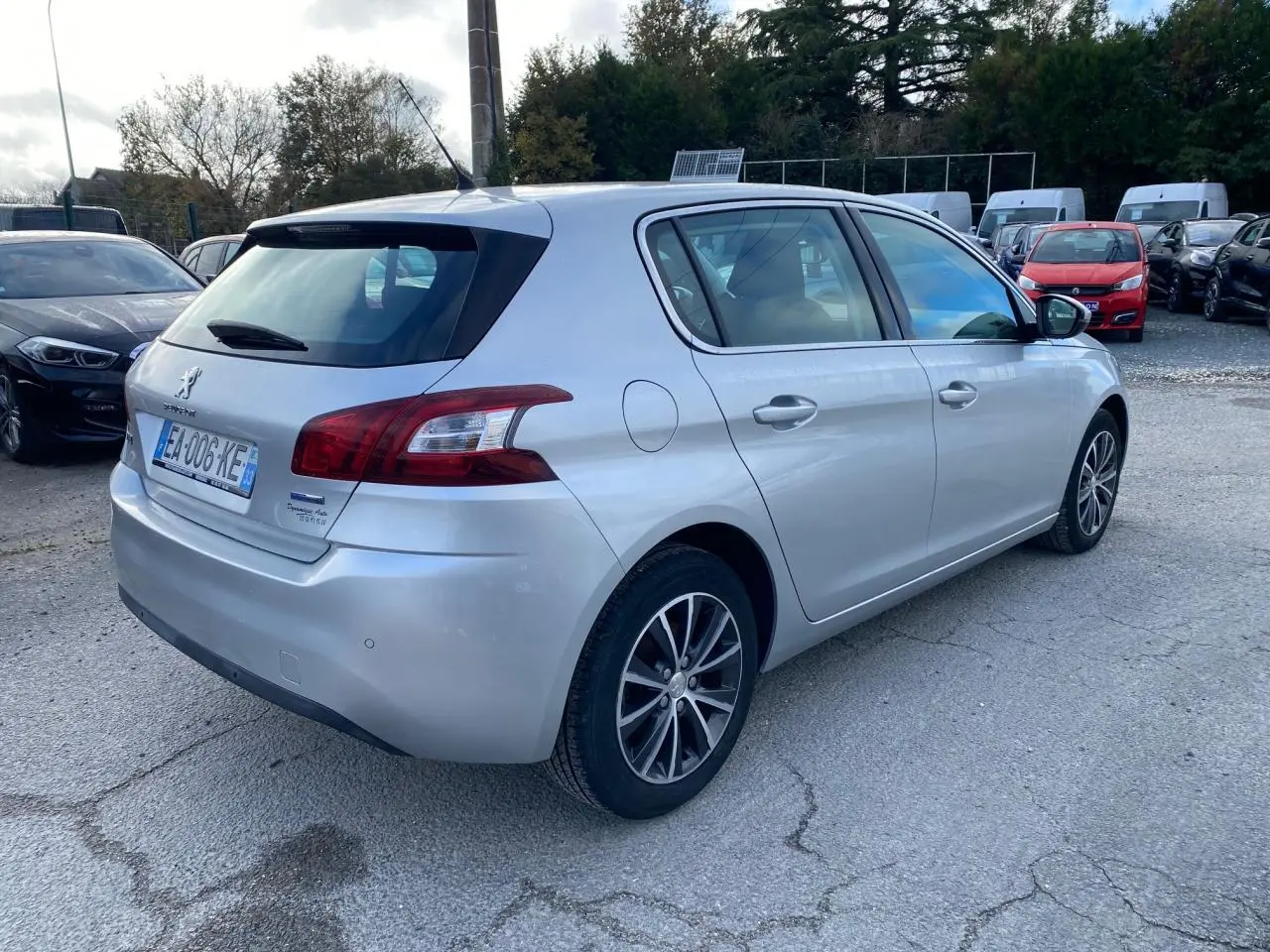Vue 3/4 arrière droite d'une Peugeot 308 gris aluminium garée sur un parking avec plusieurs autres véhicules.
