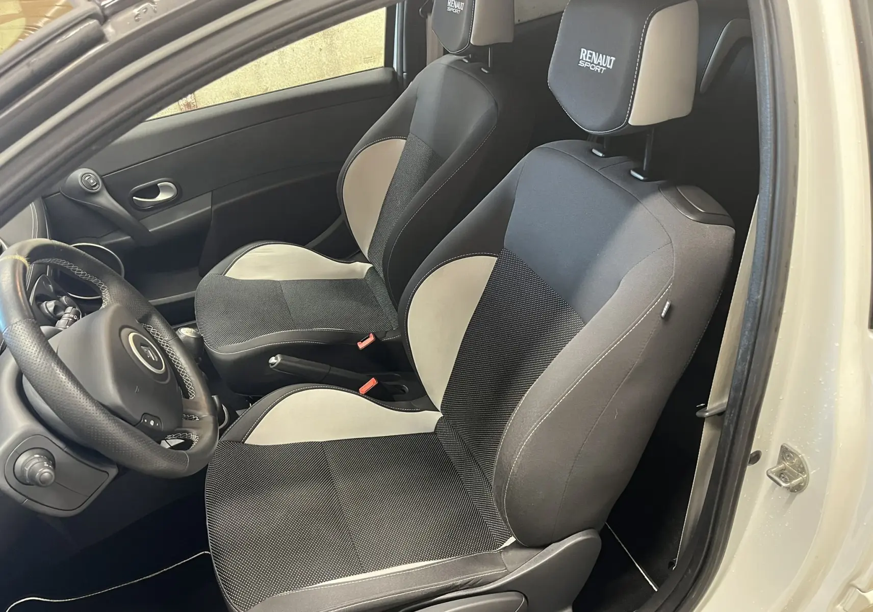 Vue intérieure côté gauche montrant les sièges avant noirs et gris avec appuie-têtes Renault Sport sur Renault Clio III blanc nacré.