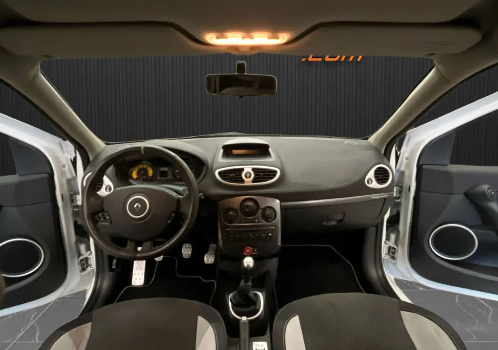 Vue intérieure avant de la Renault Clio III RS 20th Anniversary avec volant, tableau de bord noir et portes ouvertes blanc nacré.