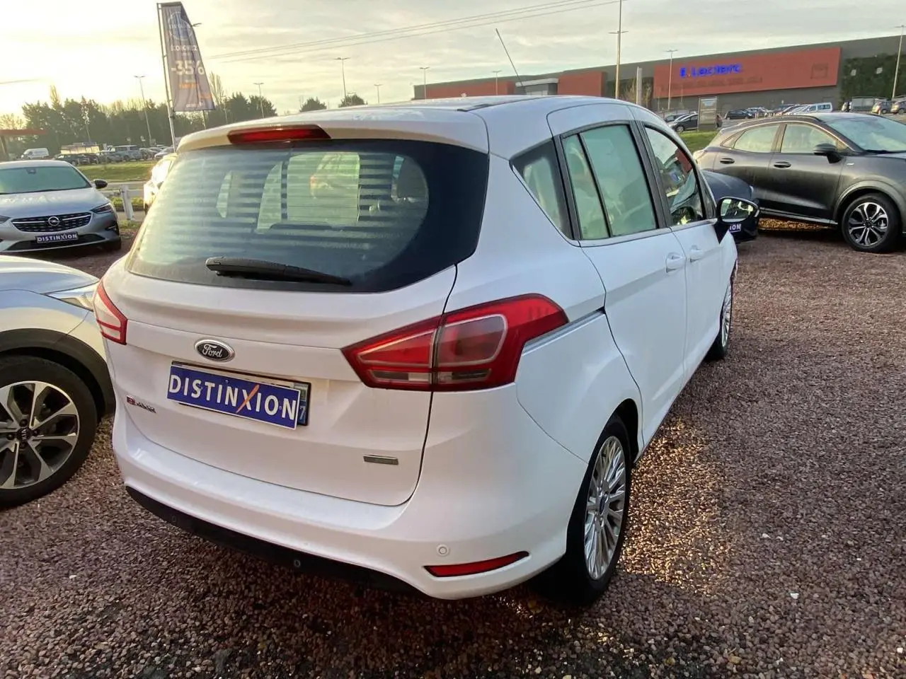 Ford B-MAX blanc vu en 3/4 arrière droit sur un parking, avec jantes alliage et feux arrière distinctifs.