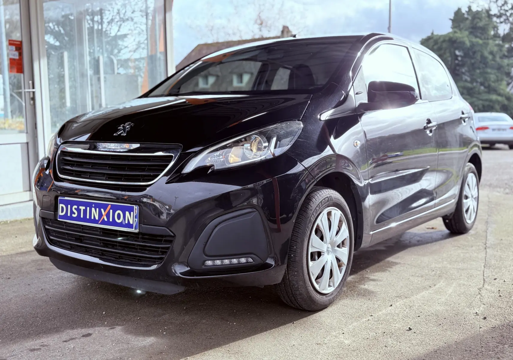 Peugeot 108 noire vue en 3/4 avant droit, avec calandre chromée et feux allumés, stationnée devant un bâtiment.