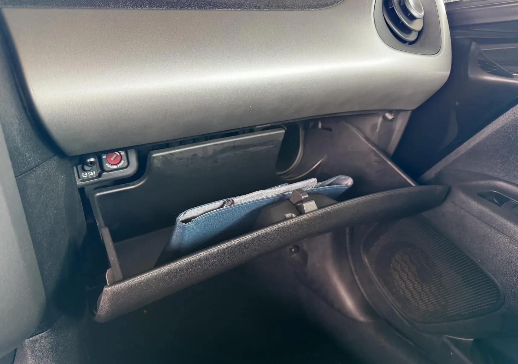 Gros plan sur la boîte à gants ouverte côté passager d'une Peugeot 108 noire, avec un livret à l'intérieur.