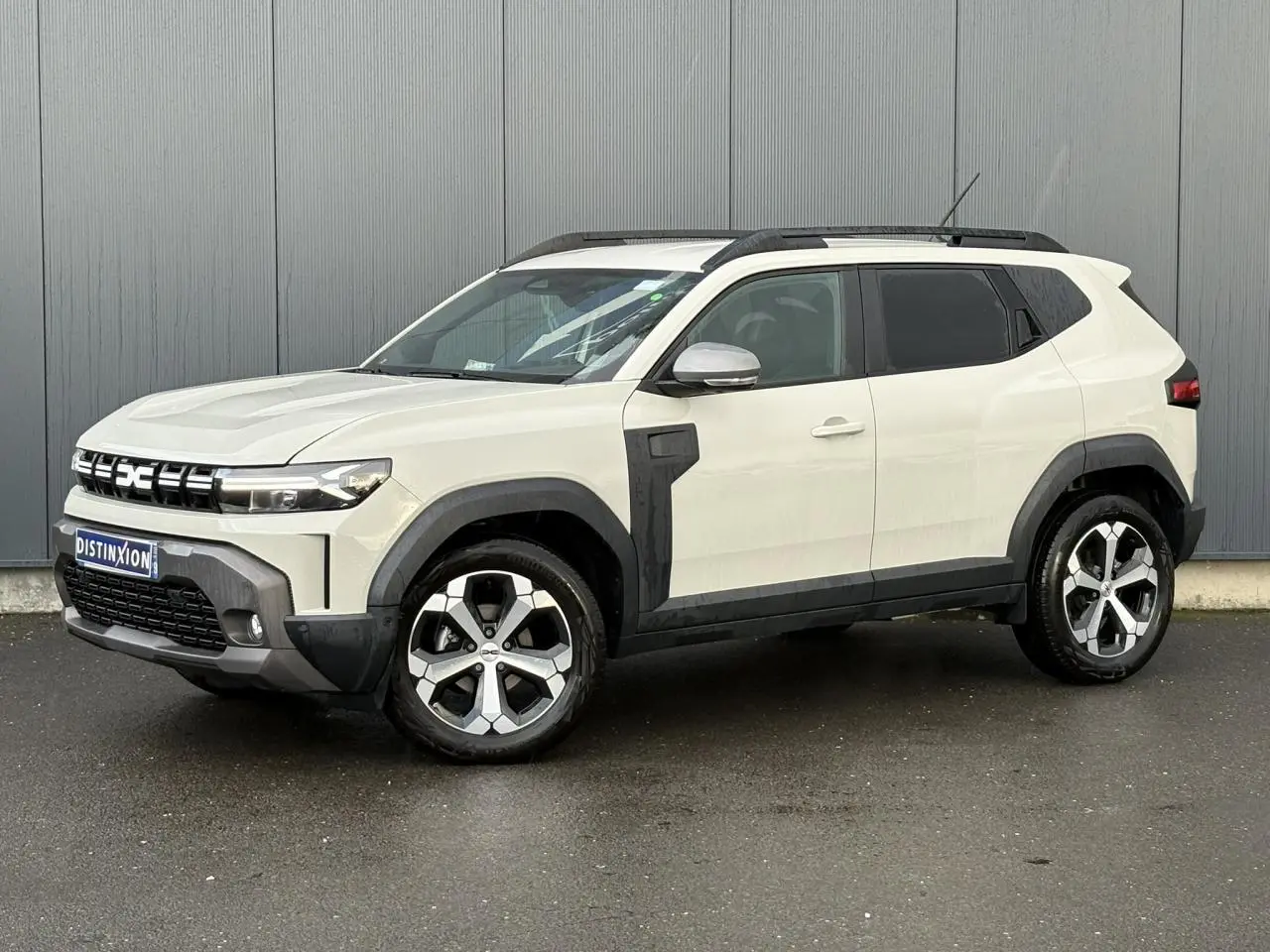 Vue 3/4 avant droit du Dacia Duster 2024 beige Sandstone avec barres de toit noires et jantes alliage distinctives.