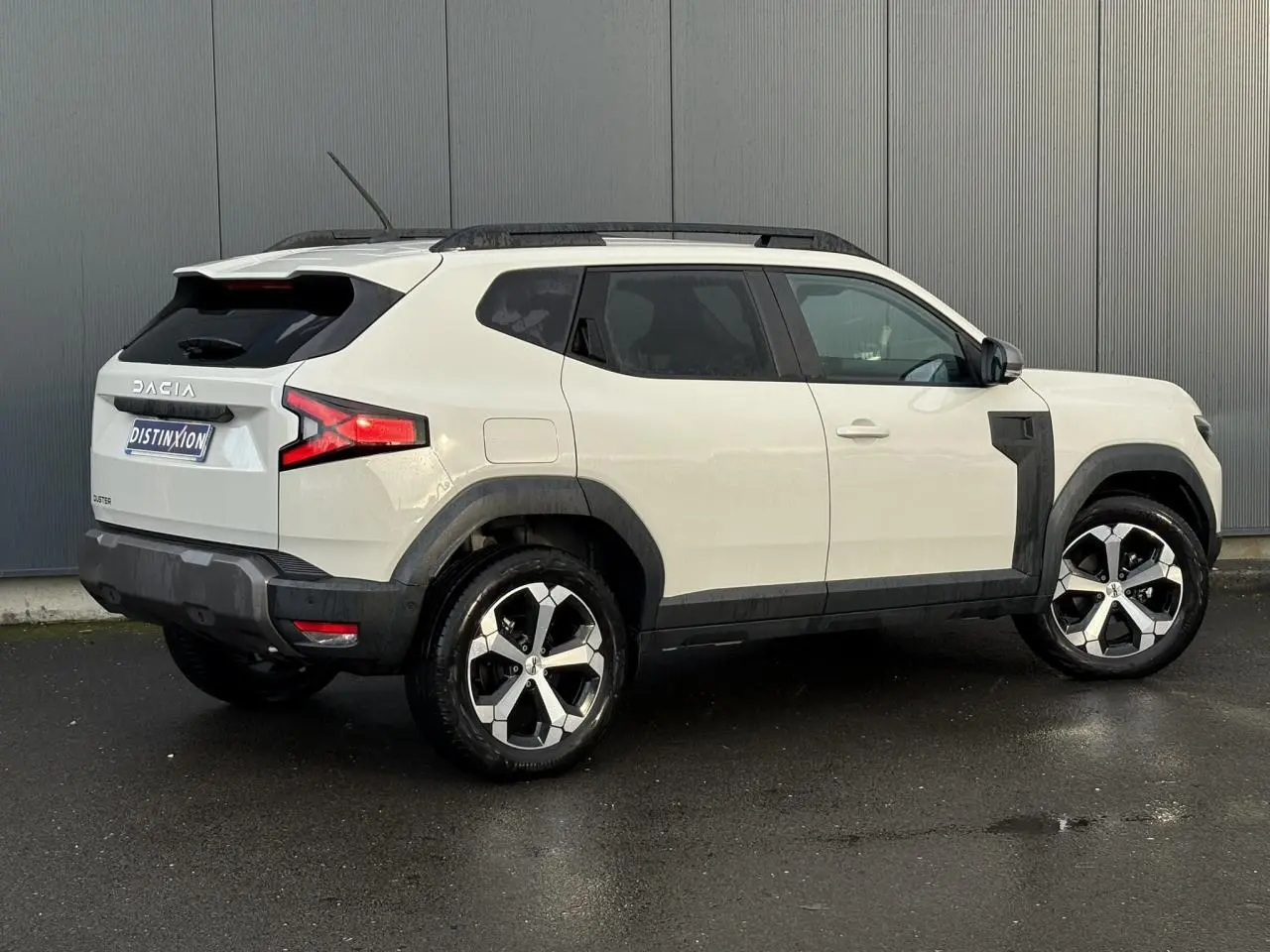 Vue 3/4 arrière droite du Dacia Duster 2024 beige Sandstone avec jantes alliage et barres de toit noires.