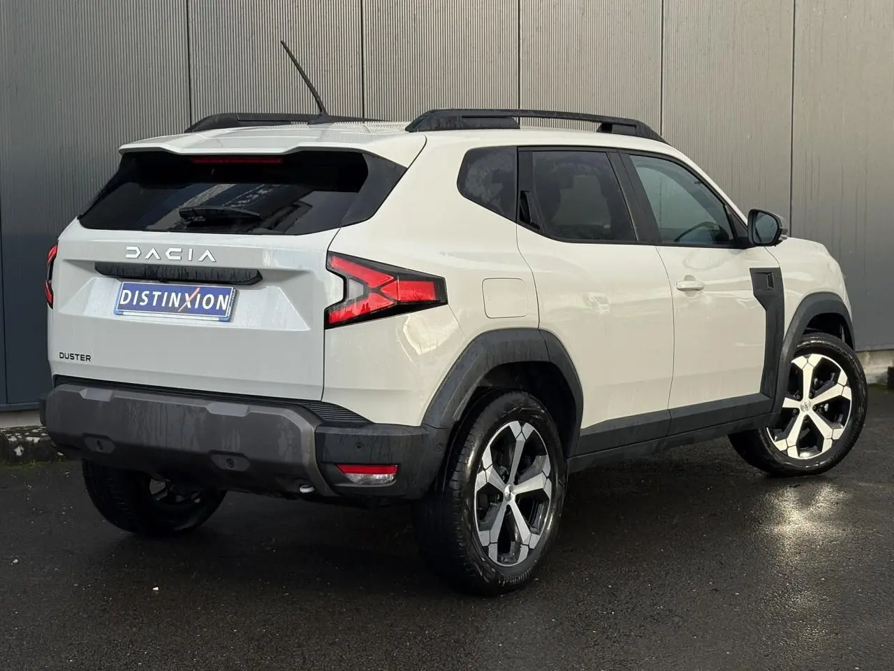 Vue 3/4 arrière droite du Dacia Duster beige Sandstone 2024 avec barres de toit noires et jantes alliage.