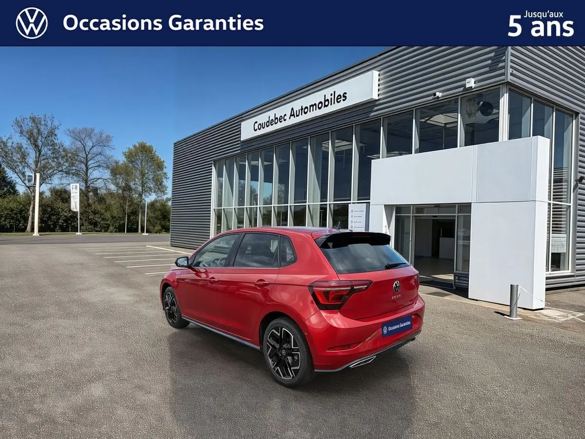 Vue 3/4 arrière droite d'une Volkswagen Polo rouge métallisée garée devant un bâtiment moderne de concession.