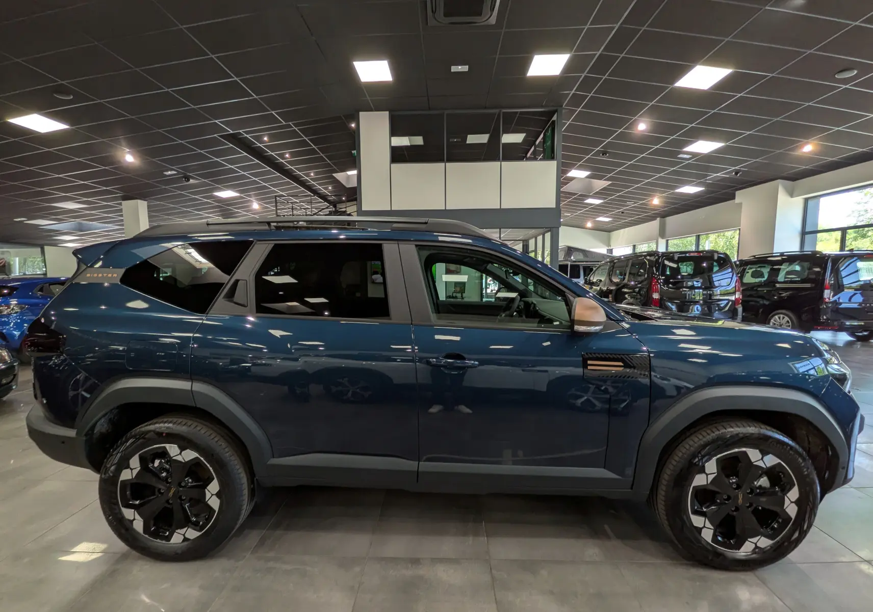 Vue de profil droit d'un Dacia Bigster bleu indigo avec jantes noires bicolores et détails noirs, en showroom.