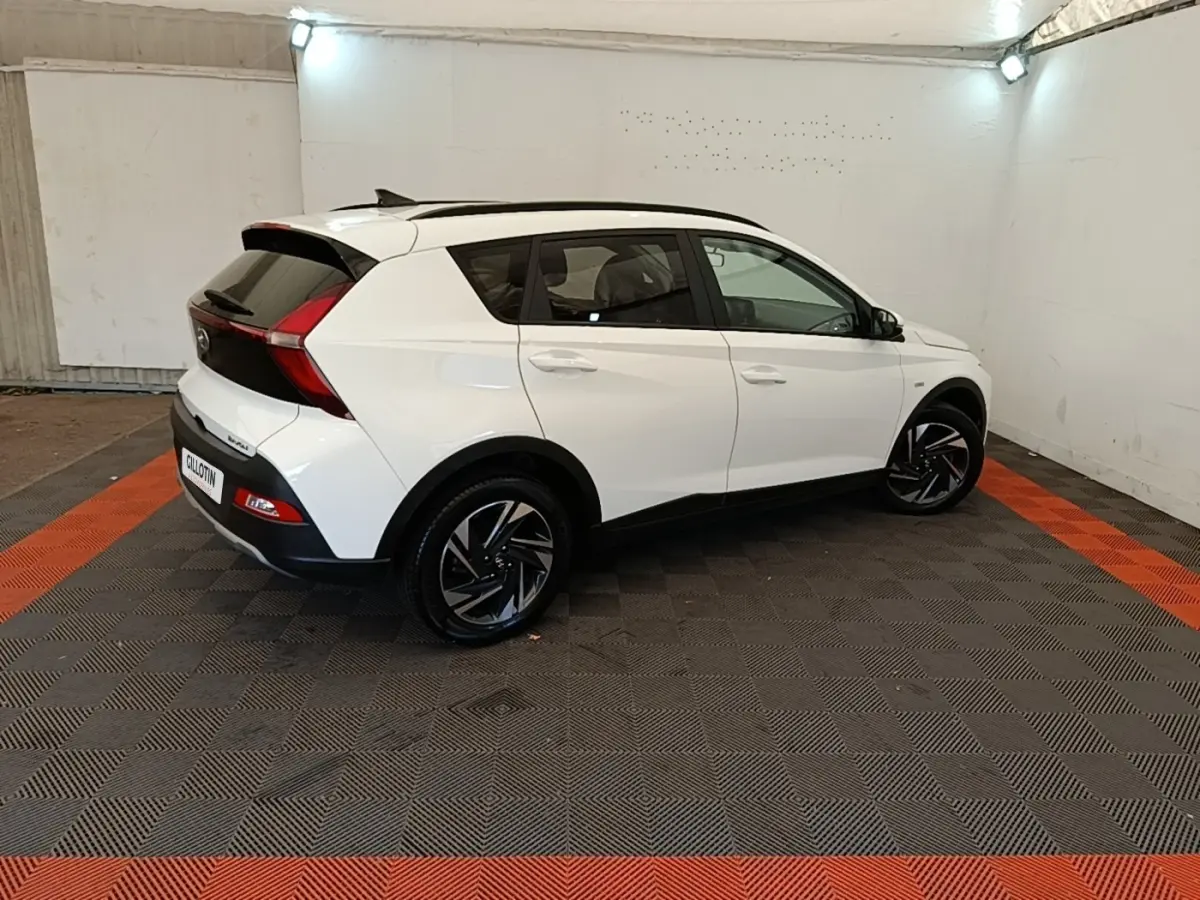 Vue 3/4 arrière droite d'une Hyundai Bayon blanche 2024 avec jantes noires et toit noir dans un garage.