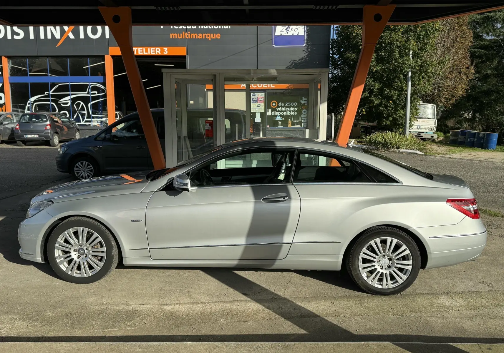 Profil côté gauche d'une Mercedes Classe E 250 Coupé gris métallisé garée devant un atelier multimarque.