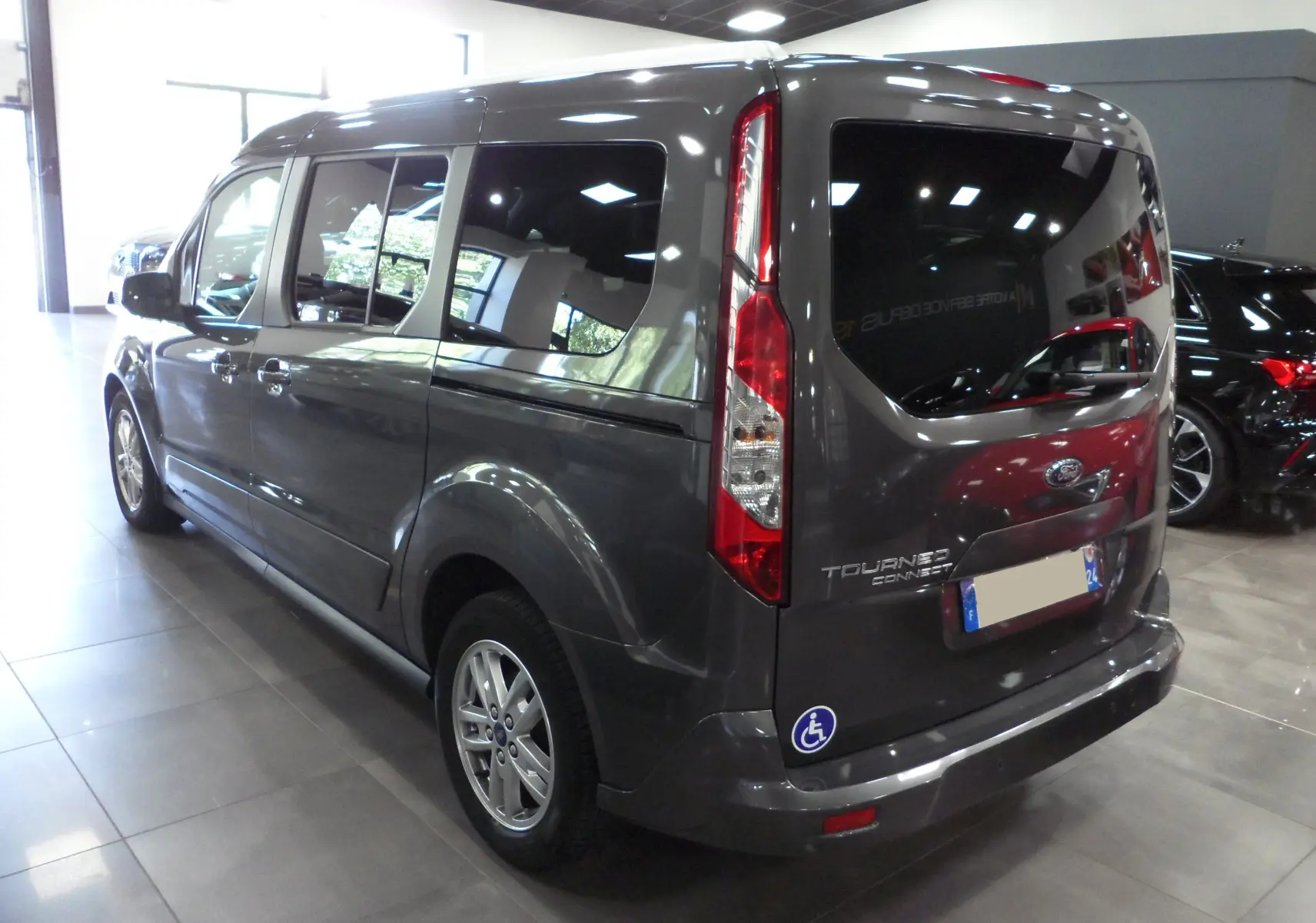 Vue 3/4 arrière droite du Ford Grand Tourneo Connect gris Magnetic avec autocollant handicapé sur le pare-chocs arrière.