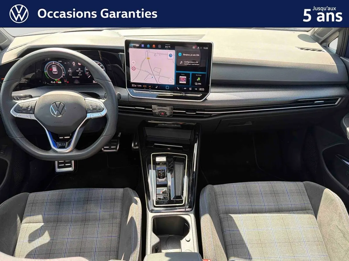 Vue intérieure avant de la Volkswagen Golf 1.5 eHybrid 2025, montrant le volant sport, l'écran tactile central et les sièges tissu à motif.