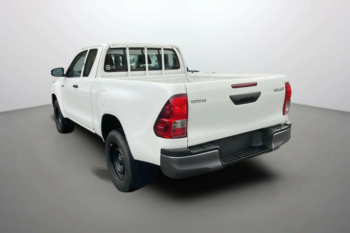 Toyota Hilux 4x4 blanc pur vu en 3/4 arrière côté gauche, avec benne ouverte et roues noires basiques.