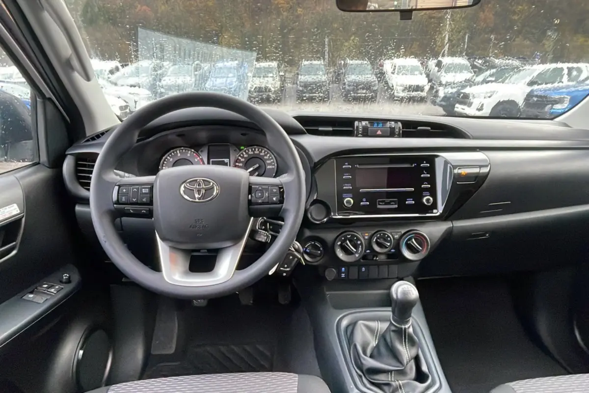 Vue intérieure avant du Toyota Hilux 2024 blanc, montrant le volant multifonction, tableau de bord et levier de vitesse manuel.