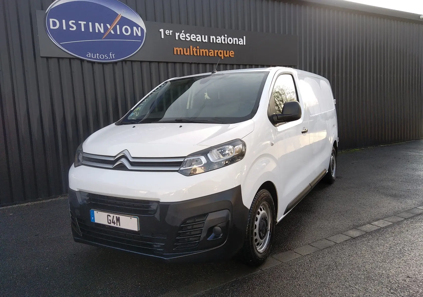 Fourgon Citroën Jumpy blanc en 3/4 avant droit, avec pare-chocs noir et jantes acier, stationné devant un mur gris.