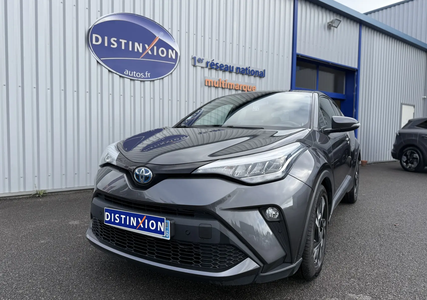 Toyota C-HR 1.8 hybrid gris atlas vu en 3/4 avant droit devant un bâtiment commercial.