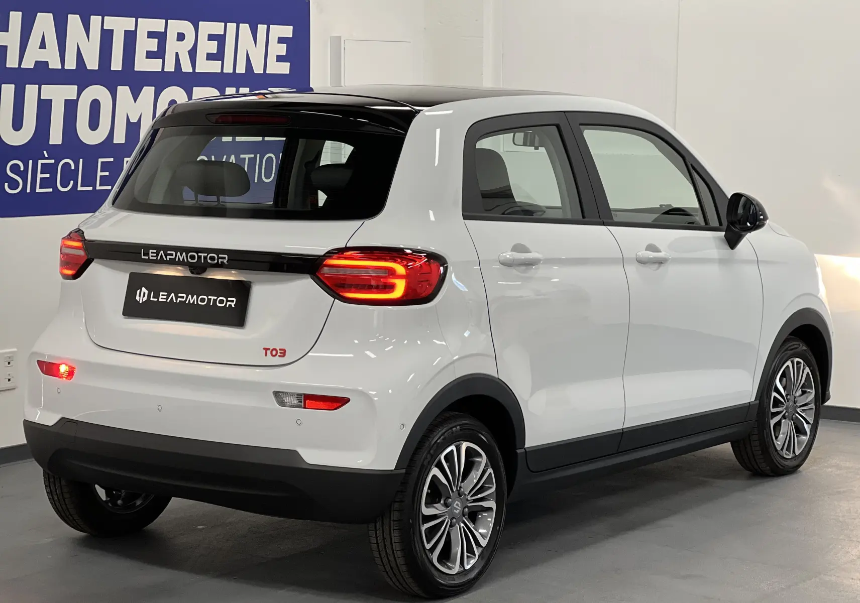 Vue 3/4 arrière droite d'une Dacia Spring blanche LEAP MOTOR T03 électrique avec feux arrière allumés en intérieur showroom.