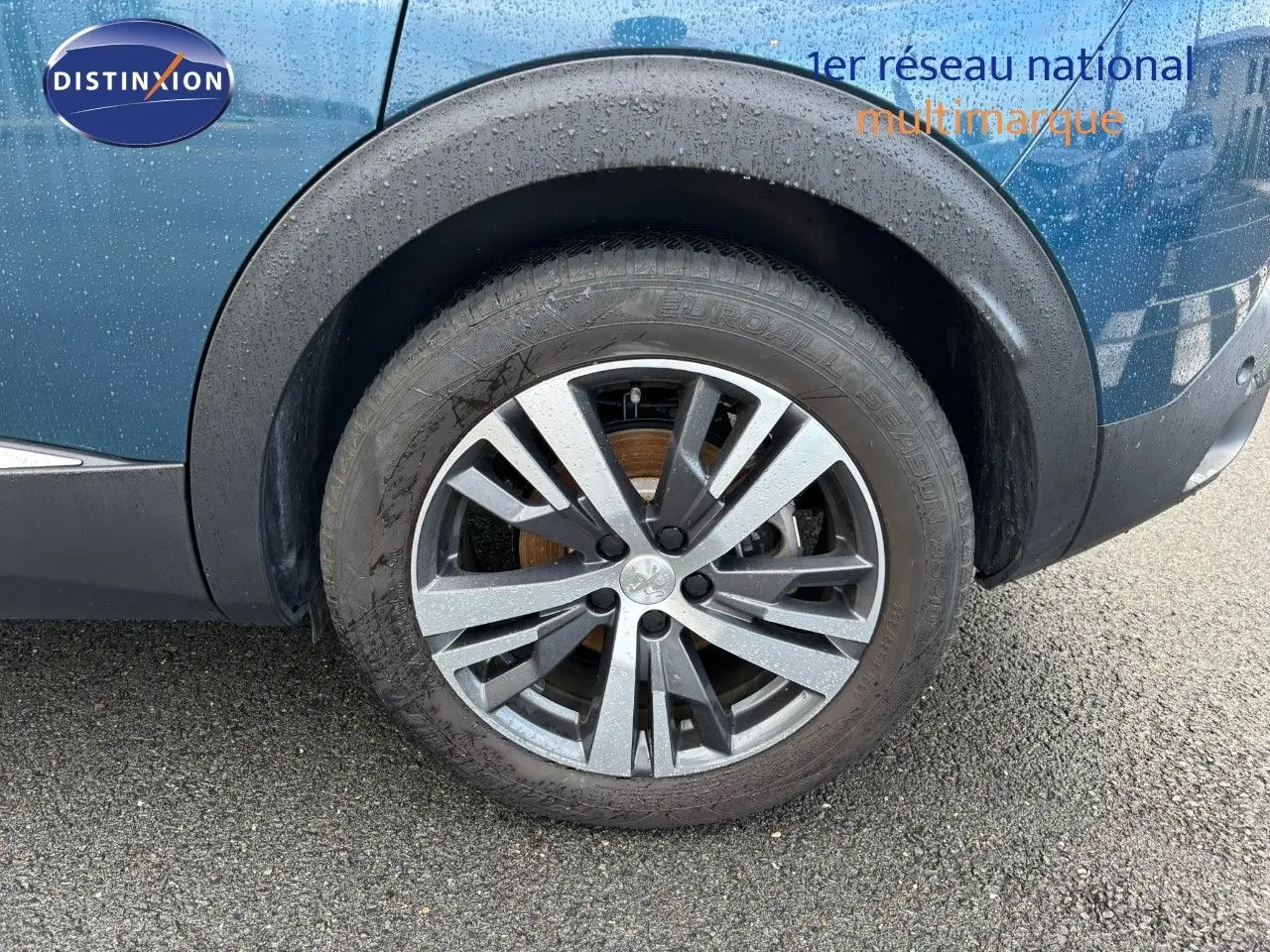 Gros plan sur la roue avant droite d'un Peugeot 3008 bleu Célèbes avec jante alliage bicolore mouillée.