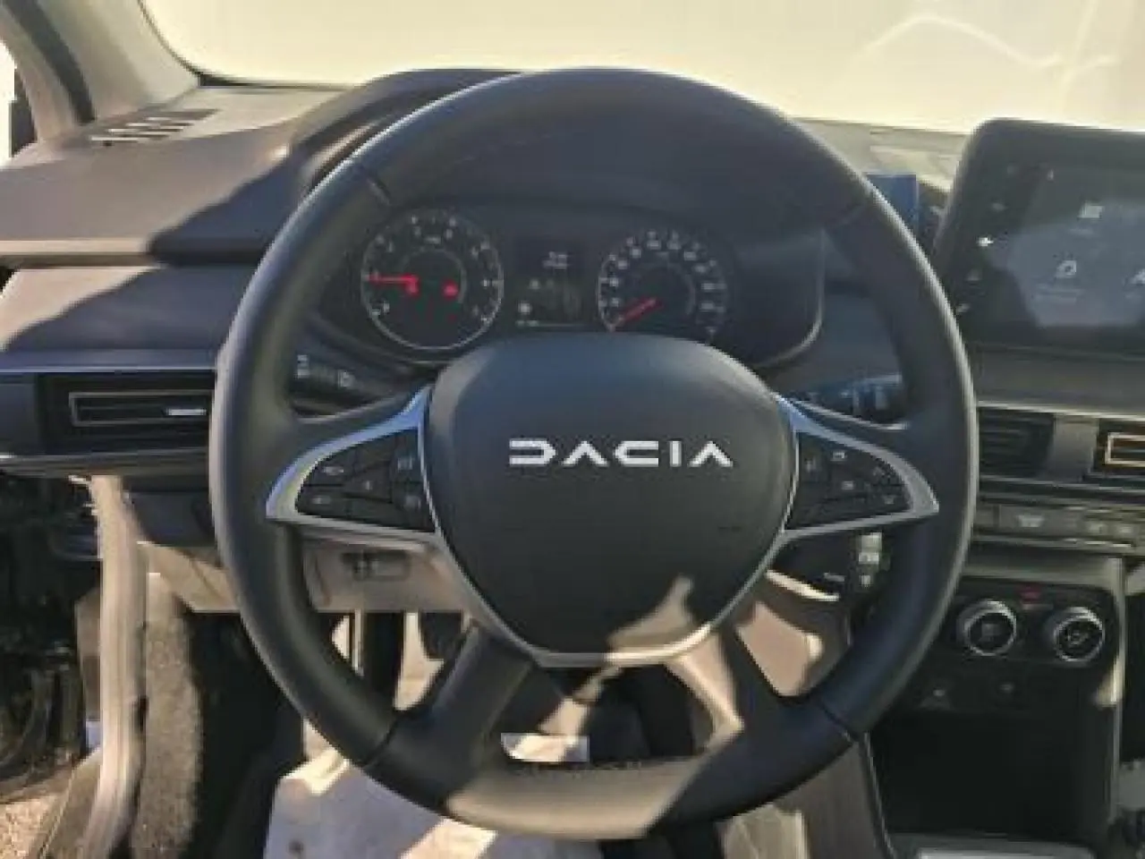 Volant noir Dacia avec commandes intégrées, tableau de bord numérique et écran tactile central visible en intérieur voiture.