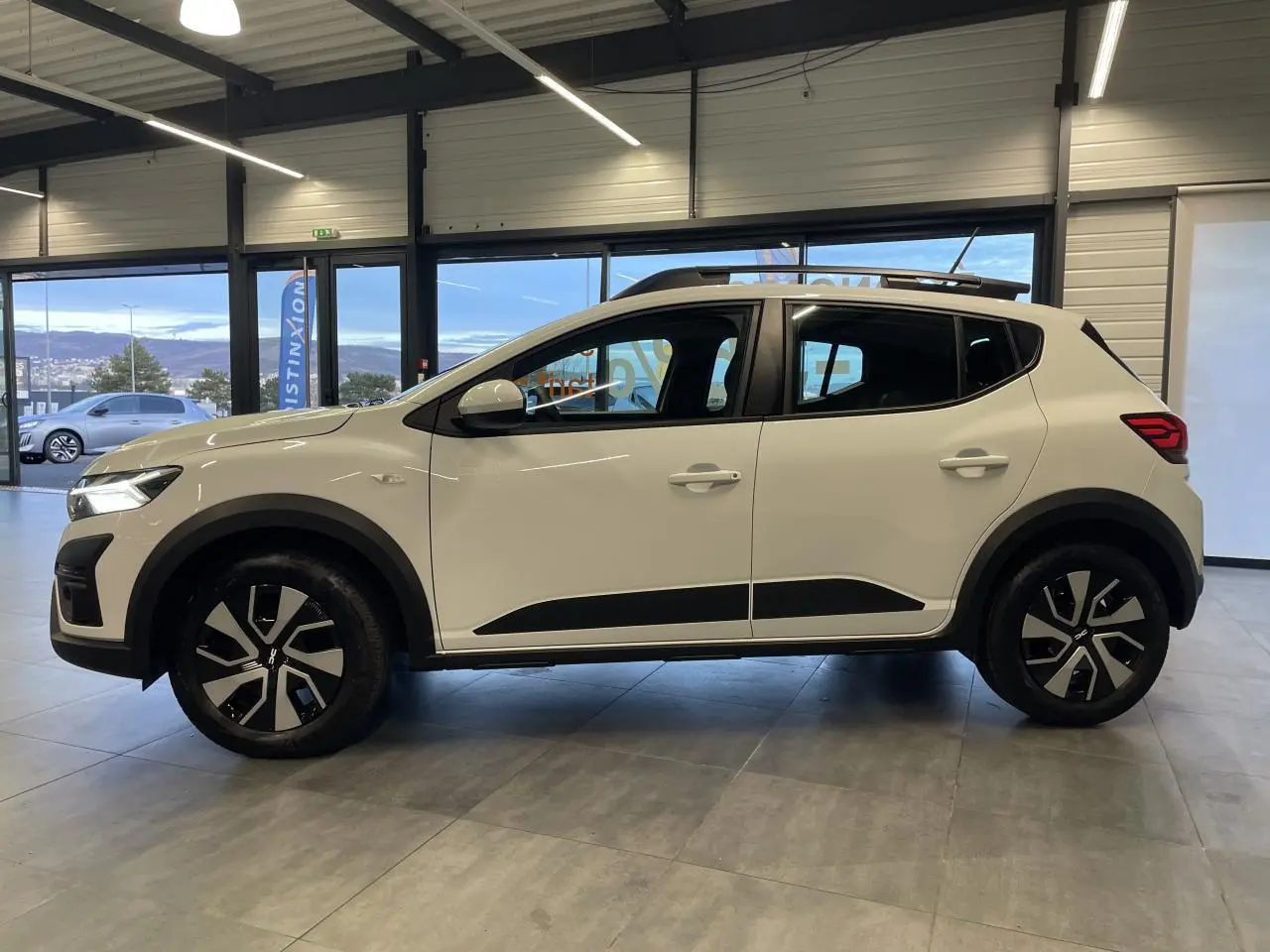 Vue latérale droite d'une Dacia Sandero TCe 90 Stepway Expression blanche Glacier avec barres de toit et jantes noires.