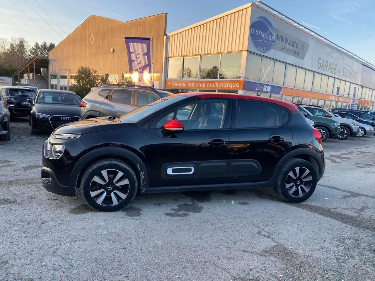 Profil côté gauche d'une Citroën C3 Shine 2023 noire avec toit et rétroviseur rouge, stationnée en extérieur.