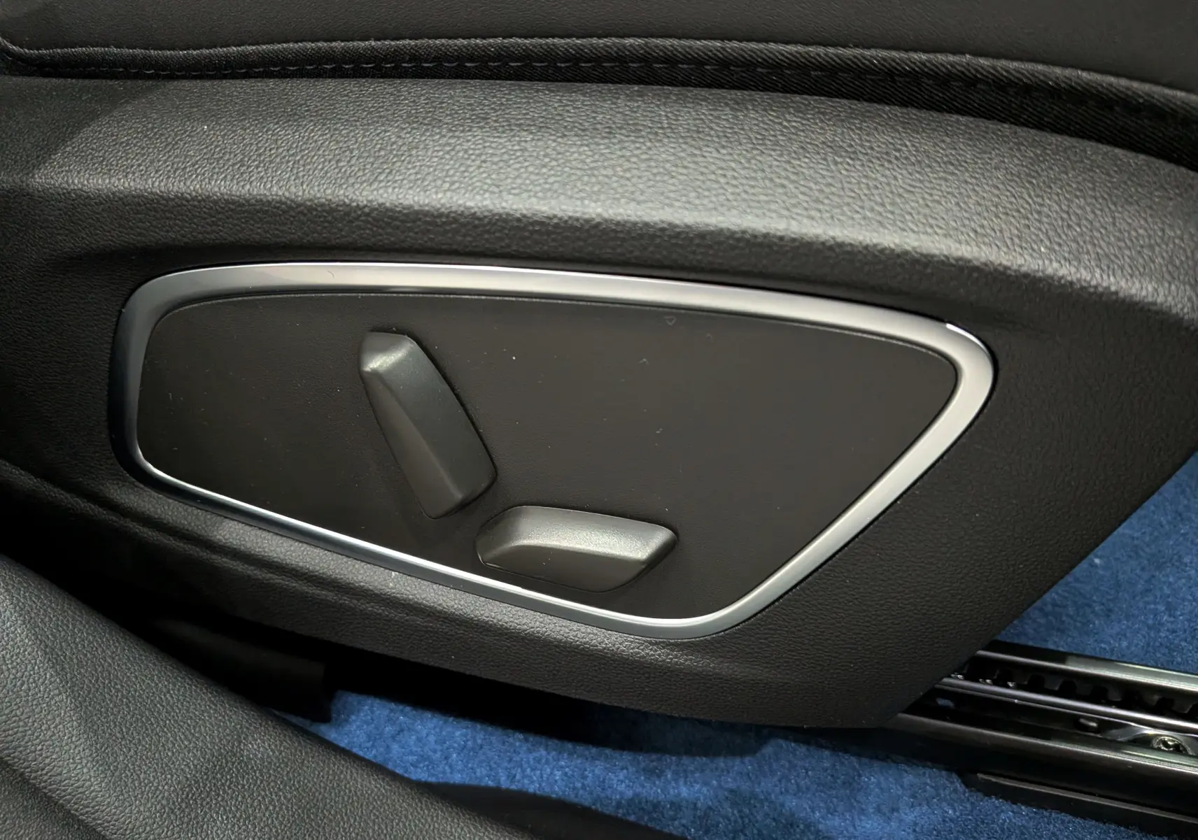 Gros plan sur les commandes électriques de réglage du siège côté gauche, intérieur noir avec moquette bleue visible.
