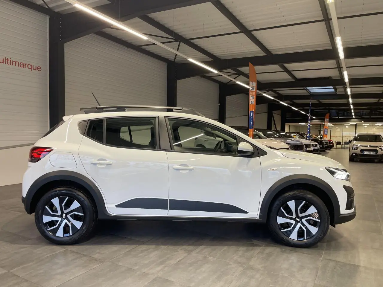 Vue de profil côté gauche d'une Dacia Sandero Stepway 2025 blanc glacier avec roues spécifiques et roue de secours visible en intérieur showroom.