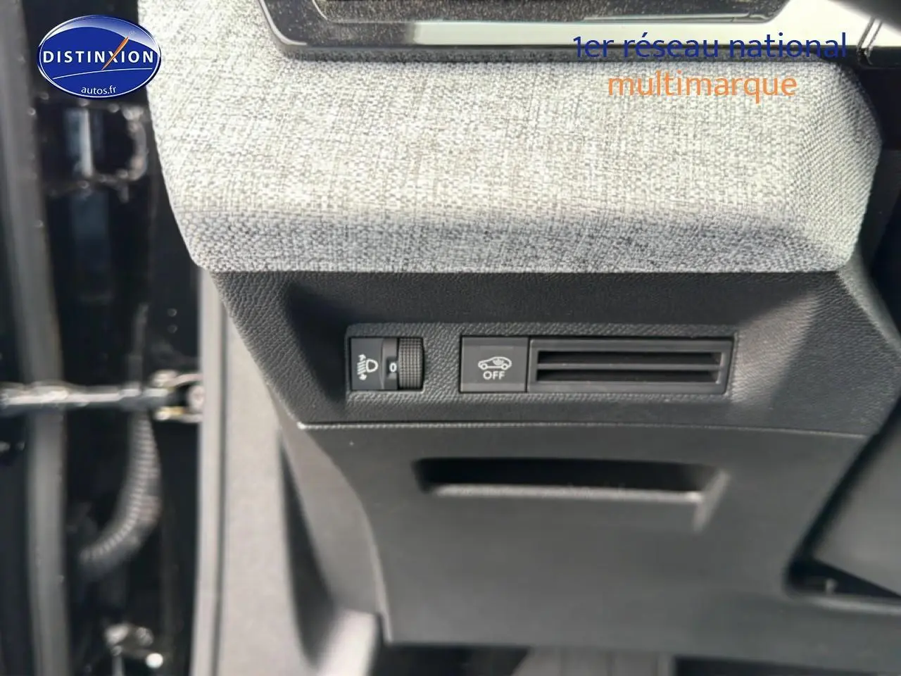 Détail intérieur montrant les commandes d'éclairage et de désactivation de l'assistance au freinage sur tissu gris et plastique noir.