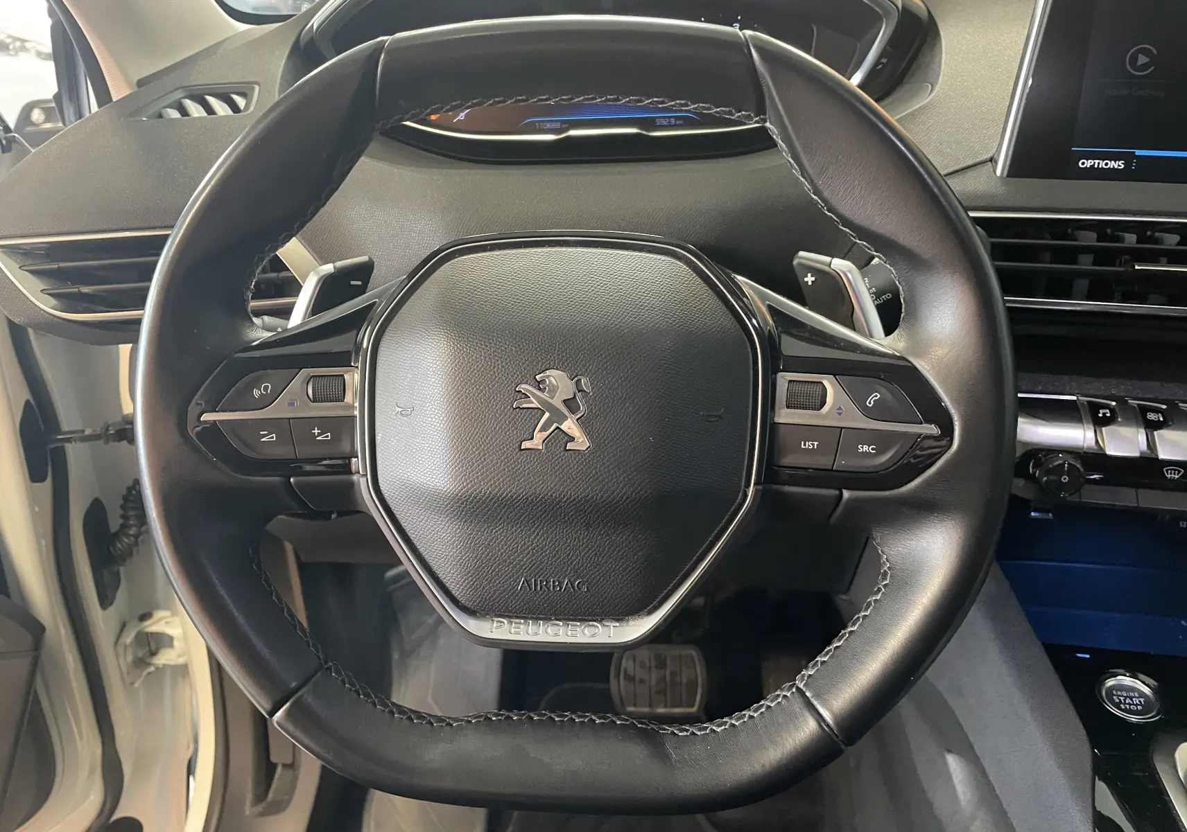 Vue rapprochée du volant cuir noir avec logo Peugeot d'un 5008 blanc nacré, tableau de bord et commandes visibles.