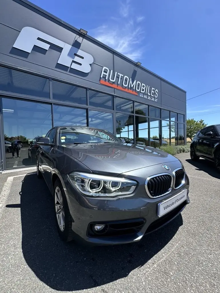 BMW Série 1 gris foncé vue en 3/4 avant droit devant un concessionnaire FB Automobiles par temps ensoleillé.