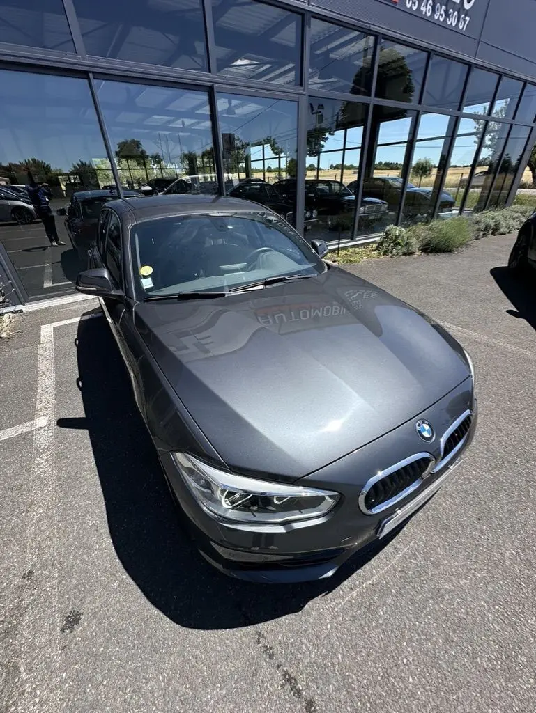 BMW Série 1 F20 gris foncé vue en 3/4 avant droit, stationnée devant une vitrine réfléchissante.