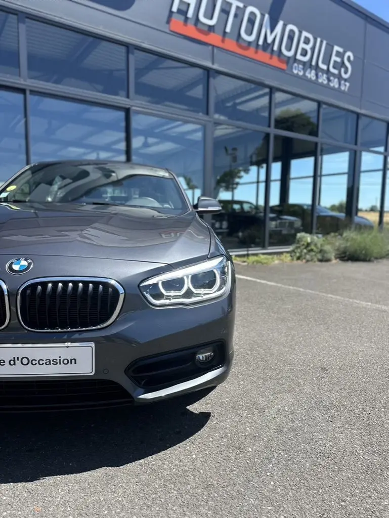 Vue avant droite d'une BMW Série 1 F20 gris foncé avec phares LED allumés devant un showroom.