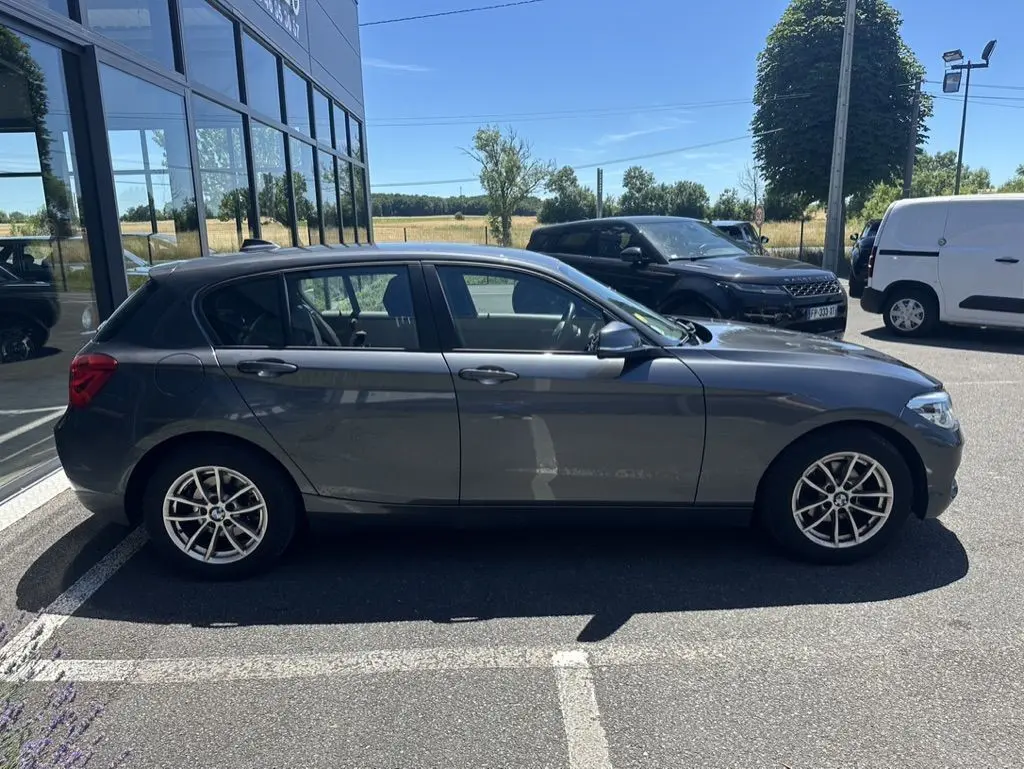 Vue de profil côté gauche d'une BMW Série 1 F20 gris foncé garée devant un bâtiment vitré par temps ensoleillé.