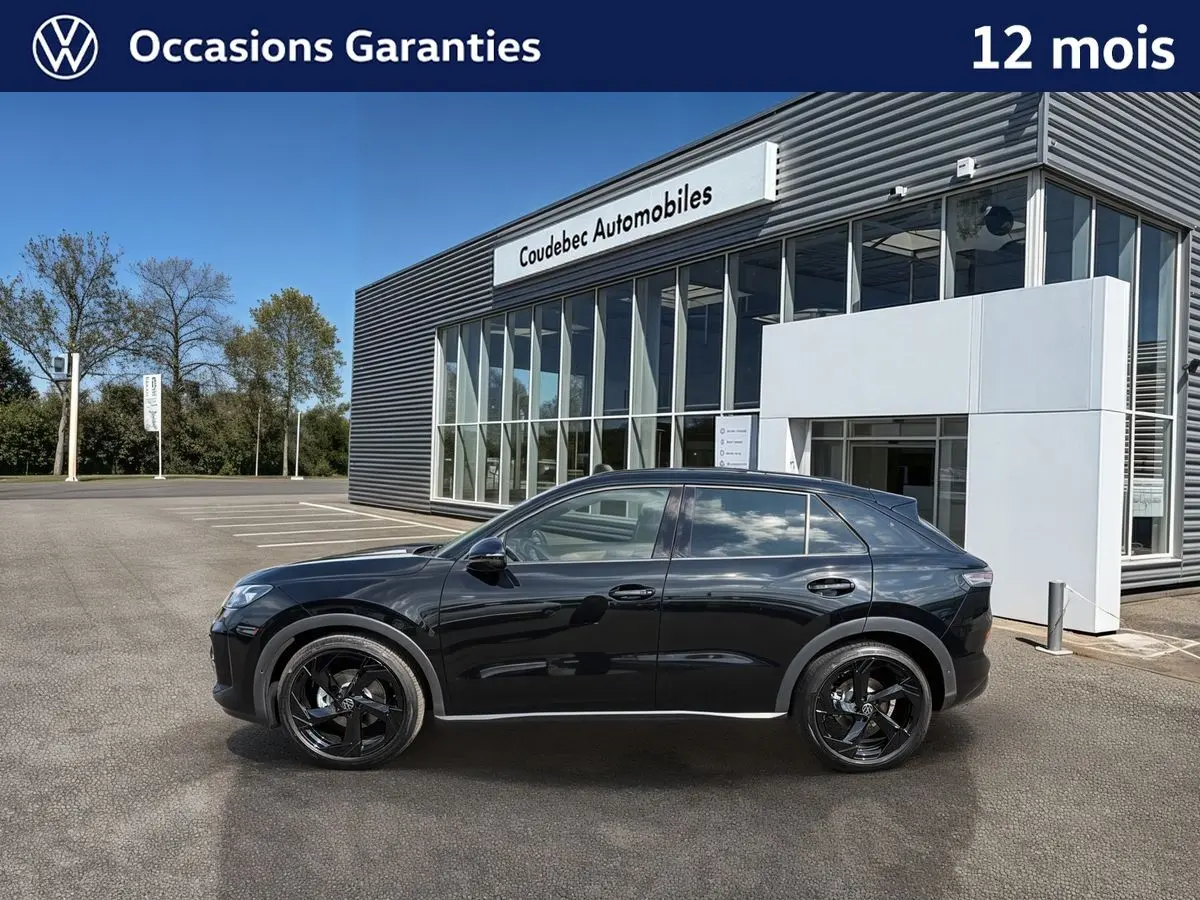Profil droit d'un Volkswagen T-Roc noir métallisé avec jantes noires, garé devant un bâtiment moderne de concession.