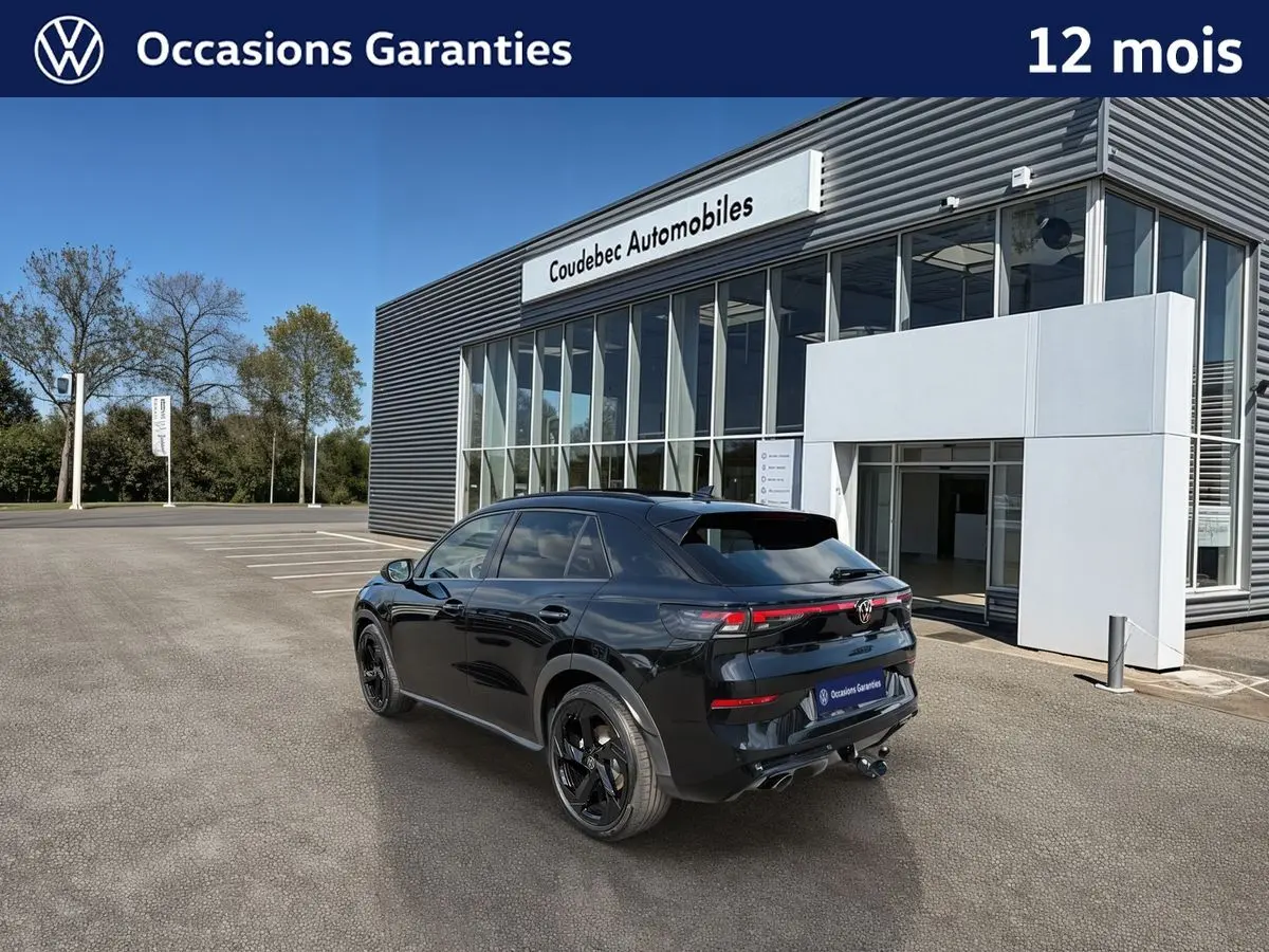 Volkswagen T-Roc noir en 3/4 arrière, avec jantes noires et crochet d'attelage, devant un bâtiment moderne.