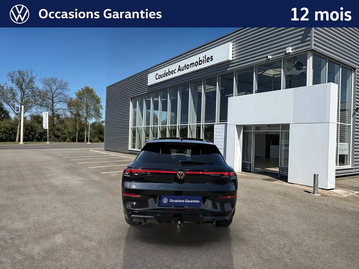 Vue arrière d'un Volkswagen T-Roc noir métallisé avec toit ouvrant panoramique et crochet d'attelage, devant un concessionnaire.