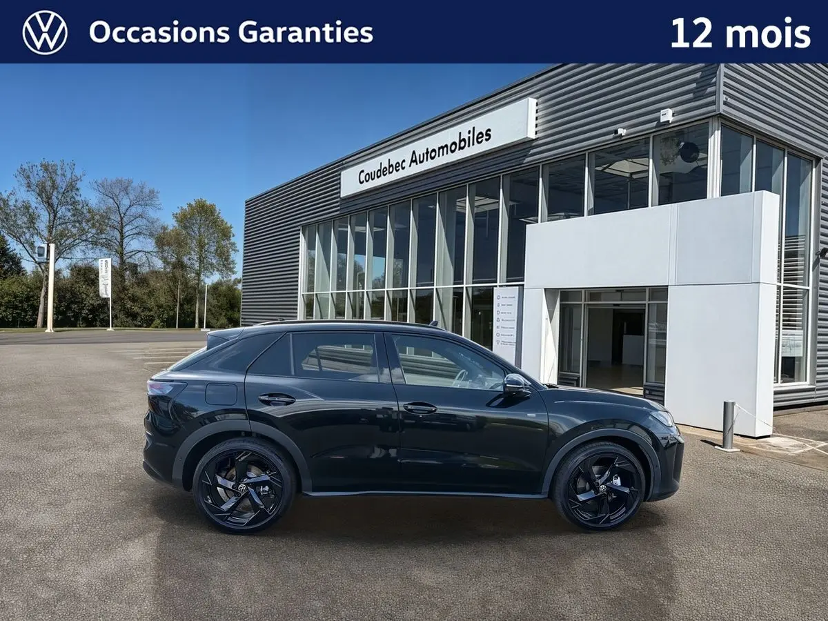 Vue de profil côté gauche d’un Volkswagen T-Roc noir métallisé avec jantes noires devant un bâtiment de concession.