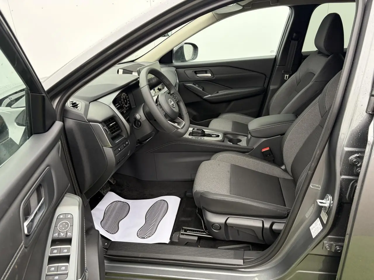 Vue intérieure côté conducteur du Nissan Qashqai III gris foncé, montrant les sièges avant et le tableau de bord moderne.