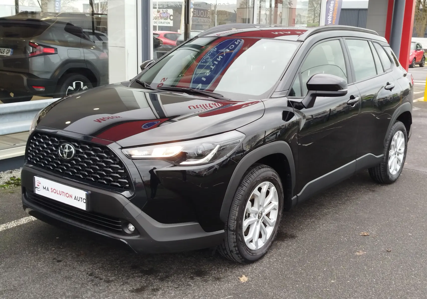 Toyota Corolla Cross noir intense vue 3/4 avant droit, avec calandre noire et jantes argentées, stationnée devant un showroom.