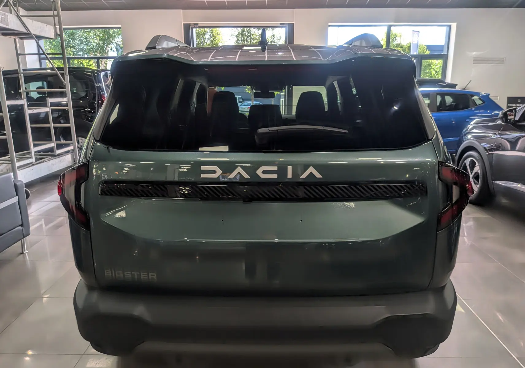 Vue arrière du Dacia Bigster vert cèdre 2026 avec logo DACIA lumineux et feux arrière sombres en intérieur showroom.