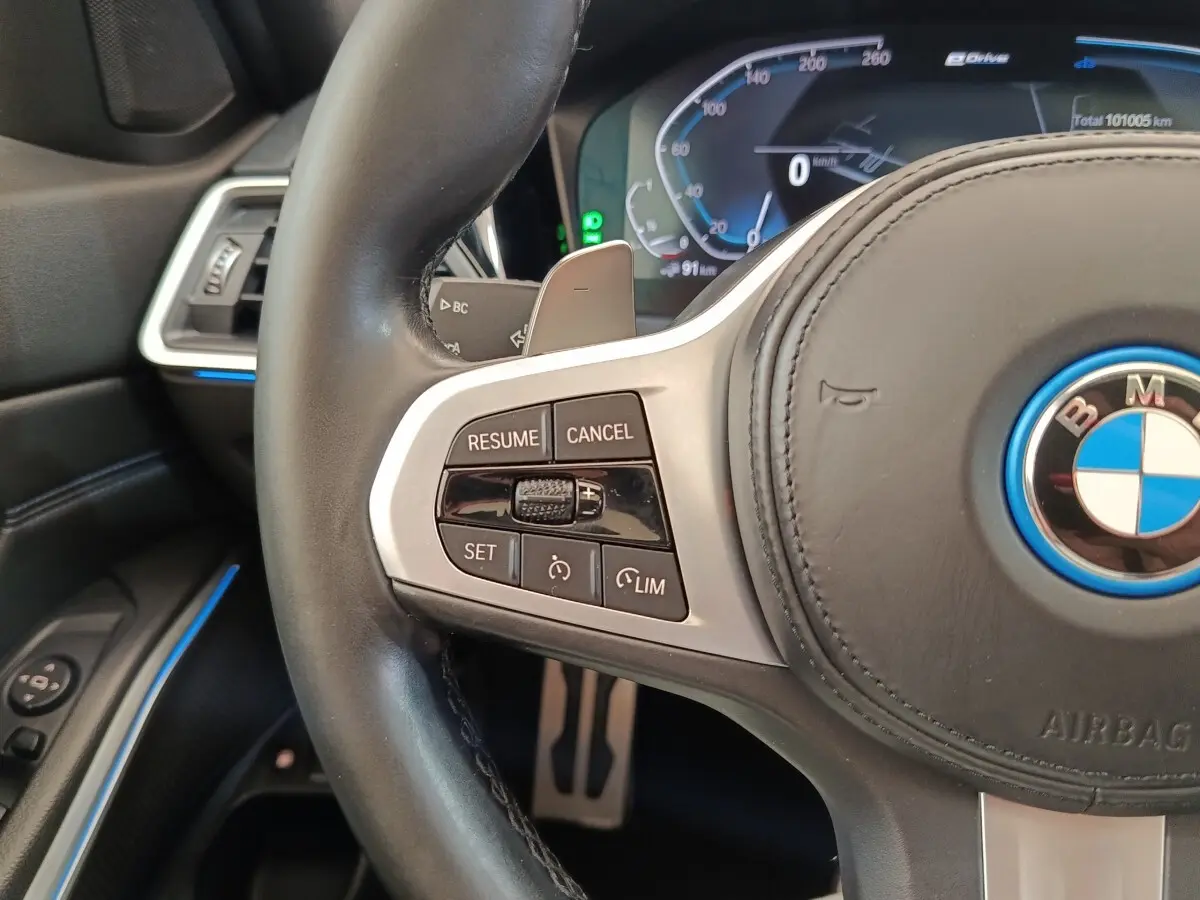Gros plan sur le volant cuir noir de la BMW Série 3 320e hybride, avec commandes de régulateur et tableau de bord numérique.