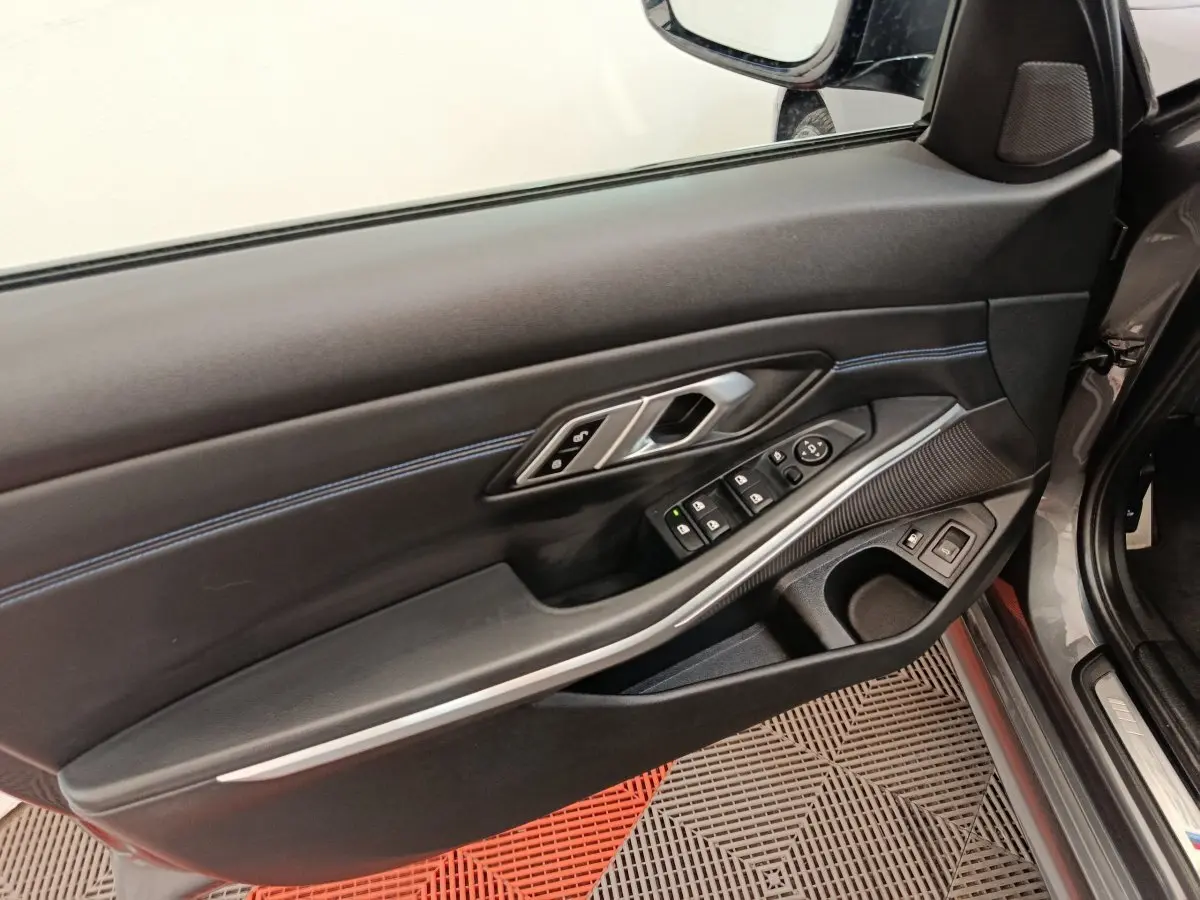 Vue intérieure côté conducteur de la porte d’une BMW Série 3 320e gris foncé avec commandes électriques et surpiqûres bleues.
