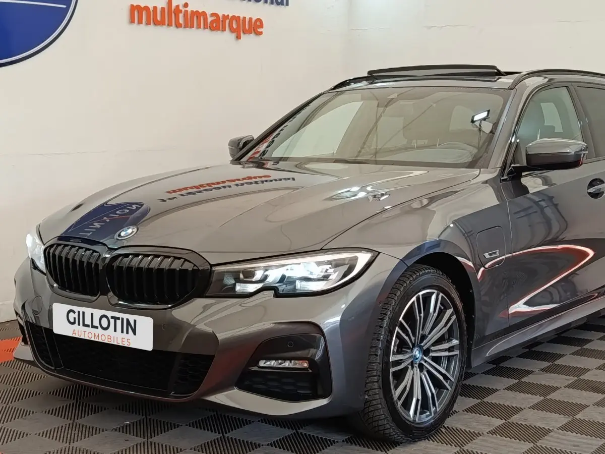BMW Série 3 320e gris foncé en 3/4 avant droit, avec phares LED allumés et jantes alliage M Sport.