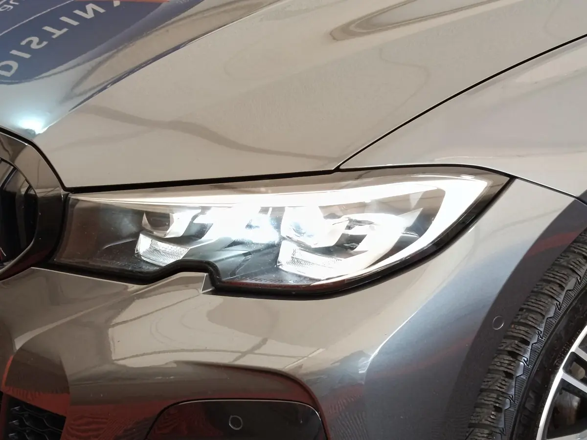 Gros plan sur le phare avant droit allumé d'une BMW Série 3 320e M Sport gris foncé, avec partie de la roue visible.