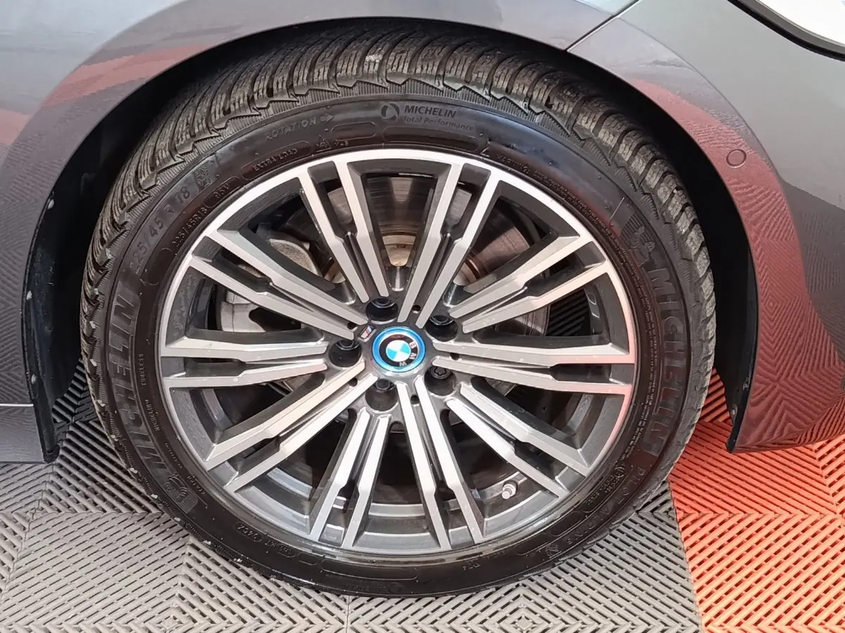 Gros plan sur la roue avant droite avec jante alliage et logo BMW, carrosserie gris foncé visible en arrière-plan.