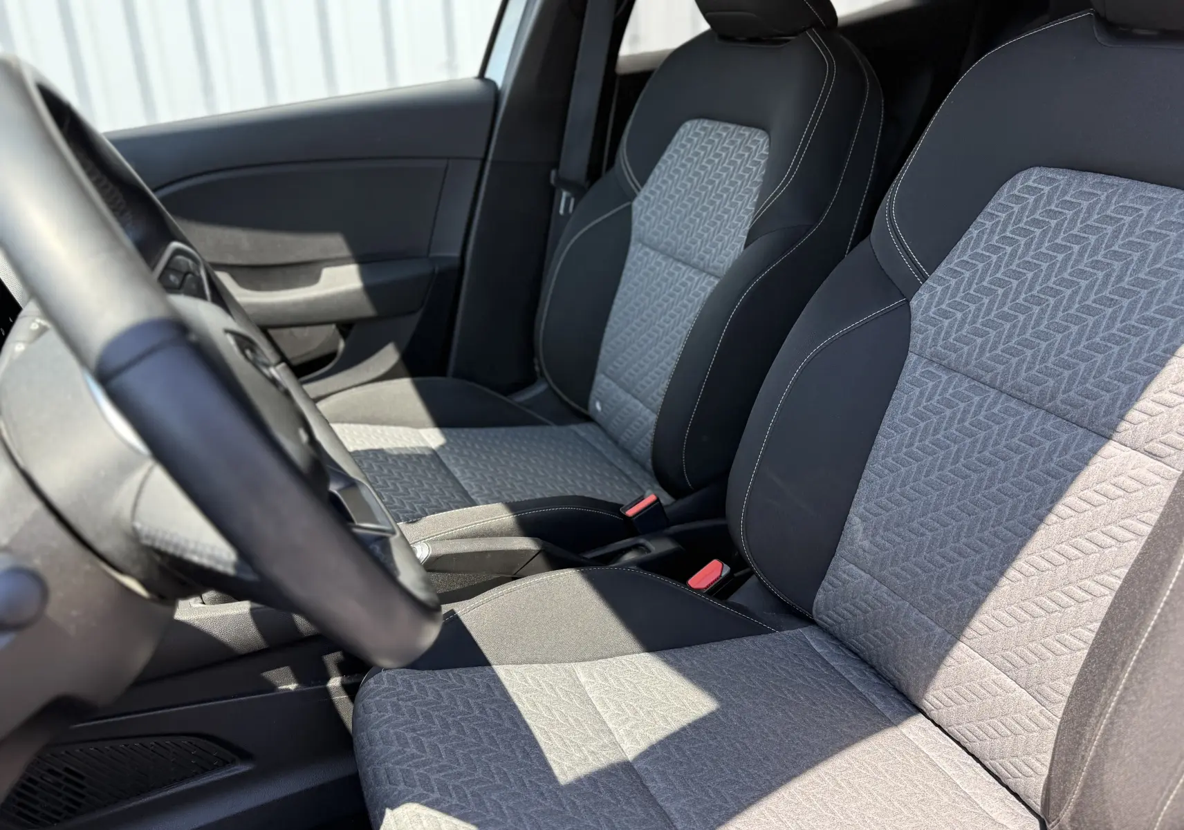 Vue intérieure côté conducteur montrant les sièges gris à motifs géométriques et le volant noir de la Renault Clio V hybride.