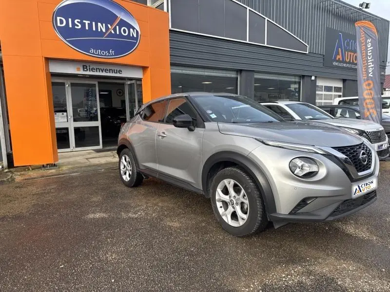 Vue 3/4 avant droit d'un Nissan Juke gris perle métallisé garé devant un concessionnaire avec logo orange Distinxion.
