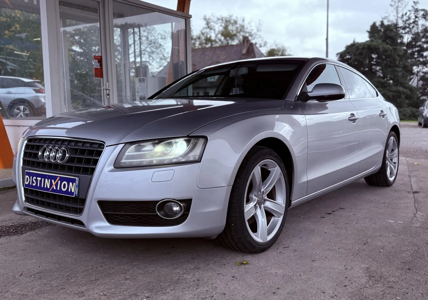 Audi A5 gris vue 3/4 avant droit avec phares allumés et jantes alliage cinq branches.