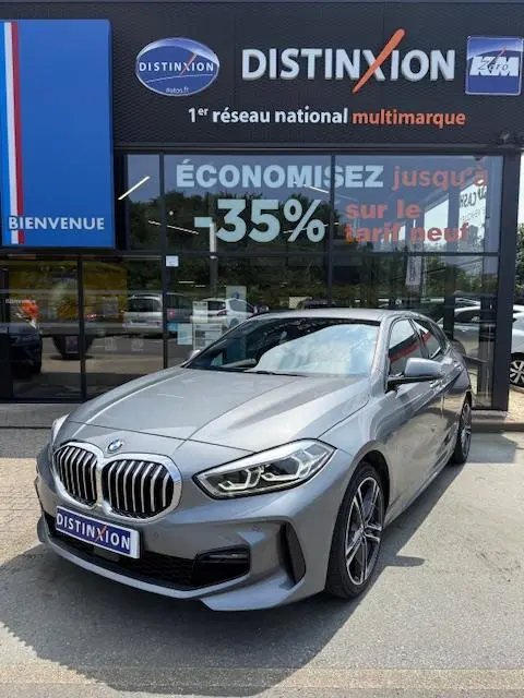 BMW Série 1 118d Edition Sport gris foncé vue 3/4 avant, avec calandre et phares LED distinctifs, devant un showroom multimarque.