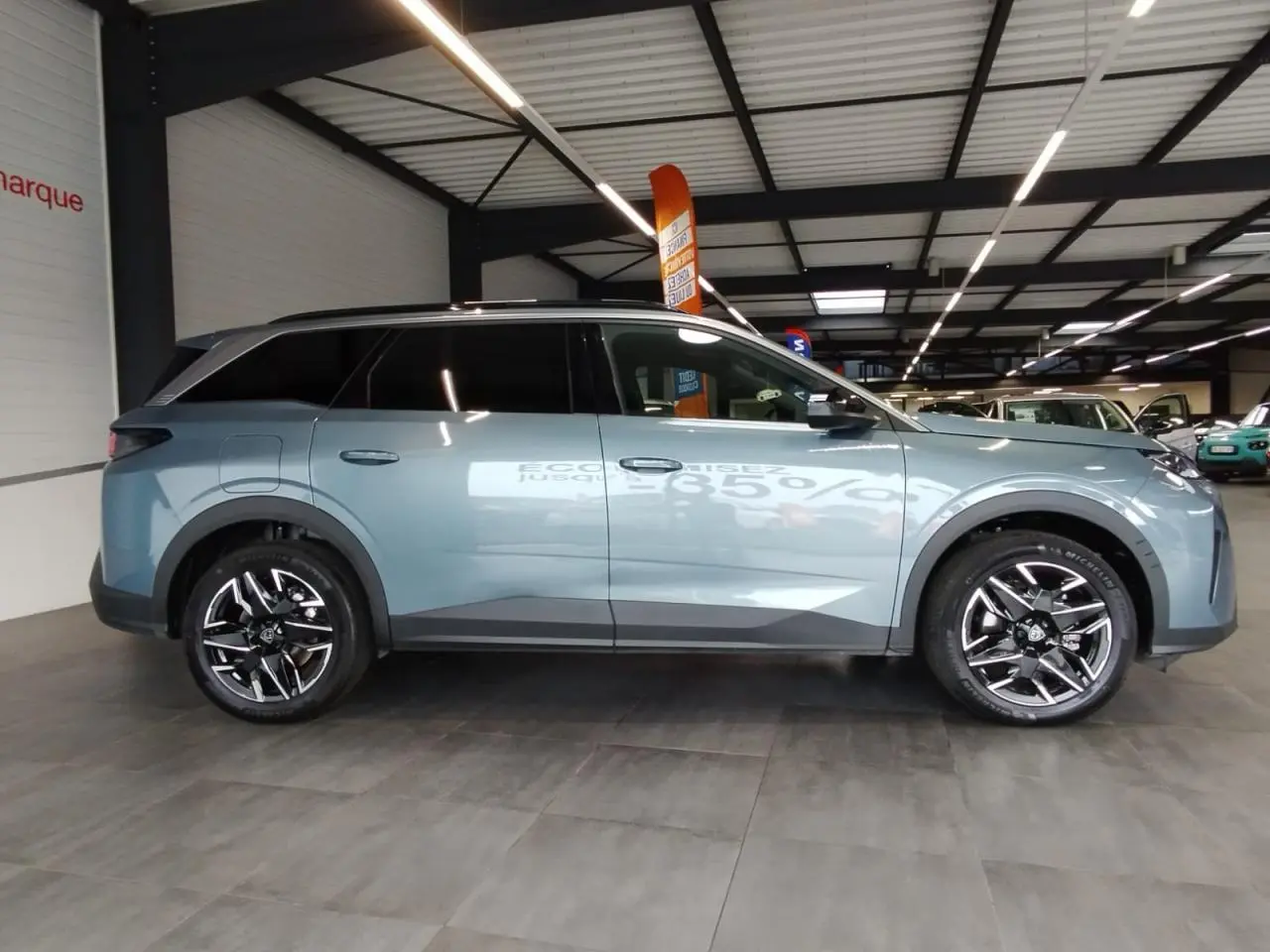Vue de profil côté gauche d'un Peugeot 5008 bleu Ingaro métallisé avec jantes alliage 19'' noires diamantées en intérieur showroom.